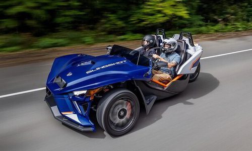 Two riders in helmets driving a blue Polaris Slingshot rental on a tree-lined mountain road